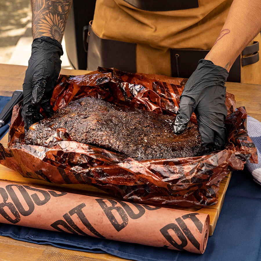 Person unwrapping a large piece of smoked meat from butcher paper on a wooden cutting board.