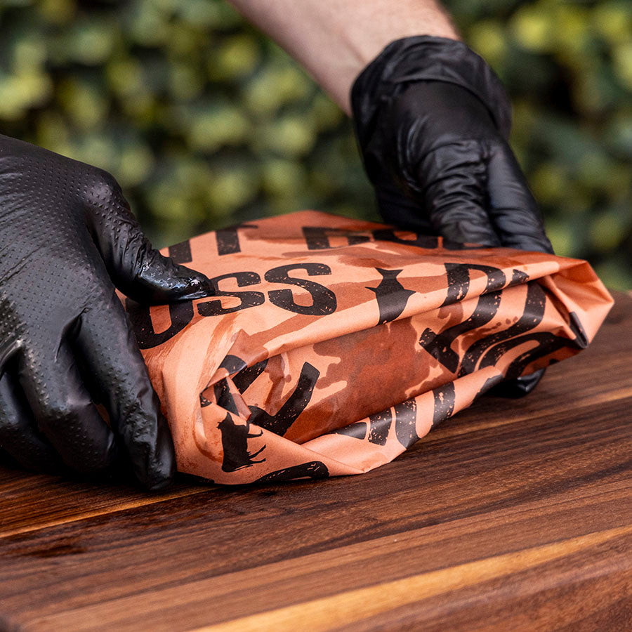 Person wearing black gloves holding meat wrapped in butcher paper on a wooden surface.